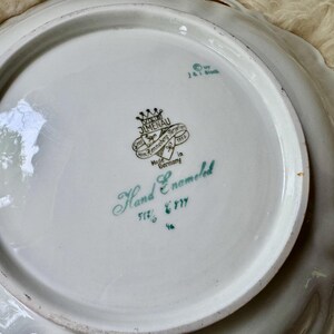 Puede incluir: Fuente de porcelana blanca con borde festoneado y dise&ntilde;o calado. El centro presenta un emblema verde y negro con las palabras "Hand Enameled" y "Made in Germany".