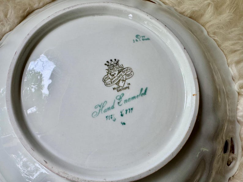 Puede incluir: Fuente de porcelana blanca con borde festoneado y dise&ntilde;o calado. El centro presenta un emblema verde y negro con las palabras "Hand Enameled" y "Made in Germany".