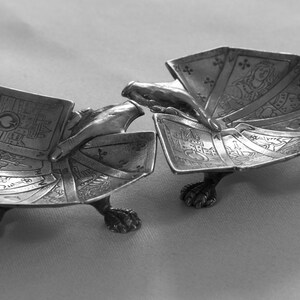 K&ouml;nnte beinhalten: Zwei silberfarbene, schildkr&ouml;tenf&ouml;rmige Aschenbecher. Jeder Aschenbecher hat eine detaillierte, f&auml;cherf&ouml;rmige Oberseite mit aufwendigen Designs und ruht auf drei Krallenf&uuml;&szlig;en. Die Aschenbecher sind offen und geben ihr Inneres frei. Der Hintergrund ist eine schlichte, hellfarbige Oberfl&auml;che.