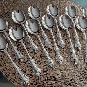 May include: A collection of silver soup spoons with ornate, floral-patterned handles. The spoons are arranged on a woven, brown placemat, reflecting light. The spoons are a classic, elegant design.