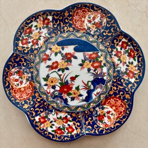 Pu&ograve; includere: Un piatto decorativo in ceramica con bordo smerlato, caratterizzato da un medaglione circolare centrale con motivi floreali rossi e gialli su sfondo bianco, circondato da un bordo blu intenso con volute dorate e motivi floreali aggiuntivi.