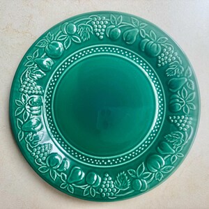 Puede incluir: Un plato redondo de cer&aacute;mica de color verde esmeralda con un centro liso y sencillo, rodeado por un borde ancho en relieve. El borde est&aacute; decorado con un patr&oacute;n repetitivo de varias frutas, incluyendo uvas, peras y hojas, con un borde punteado.