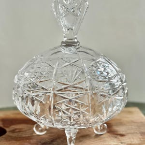 May include: A clear, round, lidded glass candy dish featuring intricate geometric cut patterns across its surface. The bowl is supported by three small, rounded glass feet and includes a matching lid with a decorative, faceted finial handle.