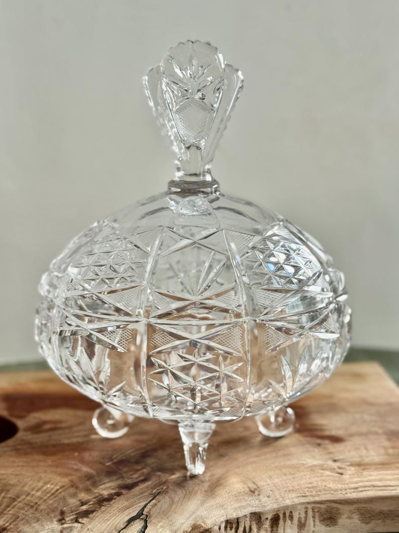May include: A clear, round, lidded glass candy dish featuring intricate geometric cut patterns across its surface. The bowl is supported by three small, rounded glass feet and includes a matching lid with a decorative, faceted finial handle.