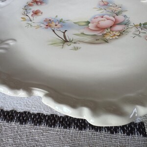 May include: A white ceramic serving platter with a scalloped edge. The platter is decorated with a floral design featuring pink and peach flowers, green leaves, and blue accents. The overall theme is vintage and decorative.