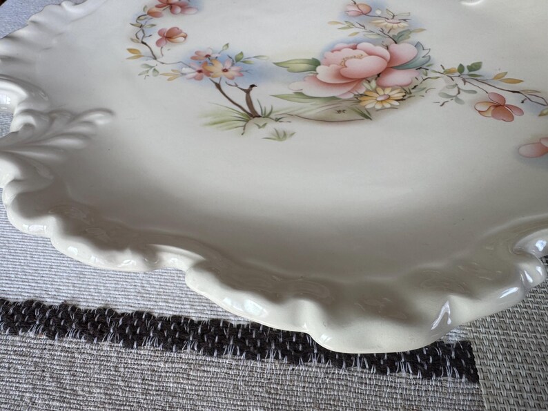 May include: A white ceramic serving platter with a scalloped edge. The platter is decorated with a floral design featuring pink and peach flowers, green leaves, and blue accents. The overall theme is vintage and decorative.