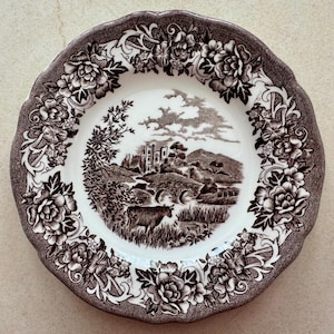 May include: A round ceramic plate with a scalloped edge featuring a dark brown transferware pattern. The center depicts a pastoral landscape with a stone bridge, a castle, and a cow. The border is decorated with detailed floral motifs.