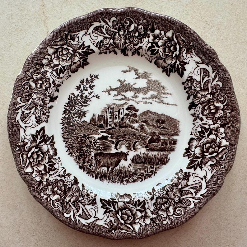 May include: A round ceramic plate with a scalloped edge featuring a dark brown transferware pattern. The center depicts a pastoral landscape with a stone bridge, a castle, and a cow. The border is decorated with detailed floral motifs.