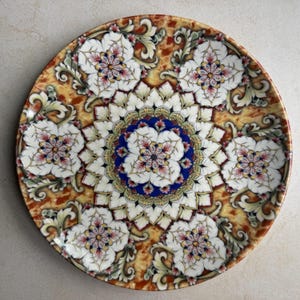 May include: A round ceramic plate featuring a central blue and white floral mandala pattern surrounded by six repeating white flower motifs. The background displays a mottled amber and brown marbled texture with intricate scrollwork details.