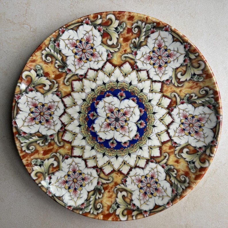 May include: A round ceramic plate featuring a central blue and white floral mandala pattern surrounded by six repeating white flower motifs. The background displays a mottled amber and brown marbled texture with intricate scrollwork details.