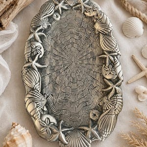 May include: An oval silver-toned metal tray featuring a detailed embossed border of various seashells and starfish. The center of the tray displays a radiating, textured pattern resembling the interior of a large scallop shell.