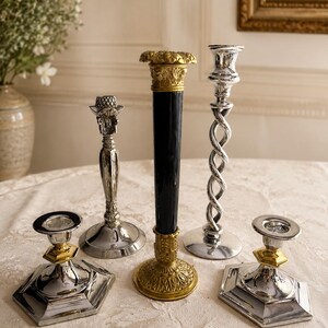 May include: A collection of five candle holders featuring two silver-toned hexagonal bases with gold accents, a silver-toned stand with a textured top, a tall black cylindrical holder with gold ornate ends, and a silver-toned twisted spiral stand.