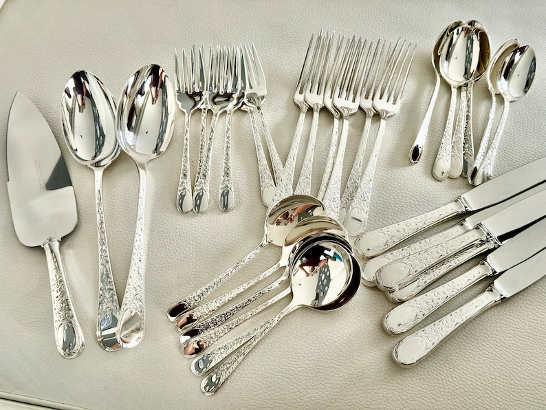 Op de afbeelding: Een set zilveren bestek, inclusief vorken, lepels en messen. Het bestek heeft een sierlijk ontwerp en is gerangschikt op een wit oppervlak.