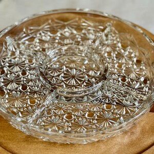 May include: Clear glass serving dish with a starburst pattern, divided into sections. The dish sits on a round wooden tray, creating a contrast in textures. Ideal for serving snacks or appetizers.