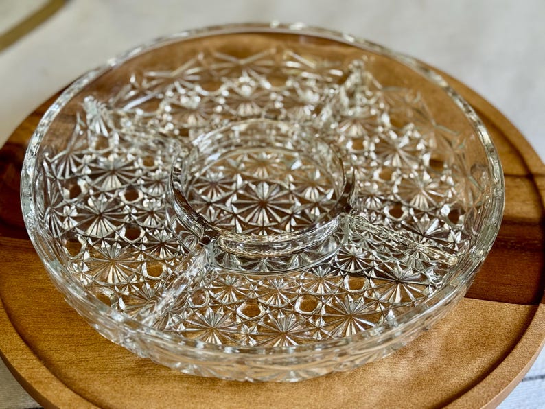 May include: Clear glass serving dish with a starburst pattern, divided into sections. The dish sits on a round wooden tray, creating a contrast in textures. Ideal for serving snacks or appetizers.