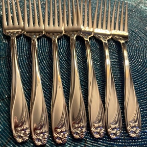 May include: A set of seven silver-toned metal forks arranged in a row. Each fork features a polished finish with a slender handle that curves slightly and terminates in a detailed, embossed floral motif at the base of the handle.