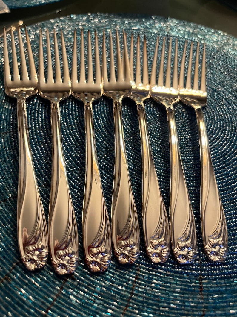 May include: A set of seven silver-toned metal forks arranged in a row. Each fork features a polished finish with a slender handle that curves slightly and terminates in a detailed, embossed floral motif at the base of the handle.