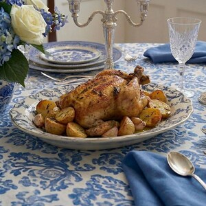K&ouml;nnte beinhalten: Ein gebratenes H&auml;hnchen und Kartoffeln werden auf einem dekorativen Teller serviert, der auf einem Tisch mit einer blau-wei&szlig;en Blumendecke steht. Auf dem Tisch befinden sich Besteck, ein Kristallweinglas und ein silberner Leuchter. Ein Strau&szlig; blauer Hortensien und wei&szlig;er Rosen befindet sich in einer Vase.