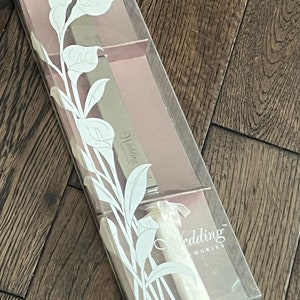 May include: A clear plastic box containing a silver cake knife with a white handle. The knife has the words "Wedding Memories" engraved on the blade. The box is decorated with a white floral design.