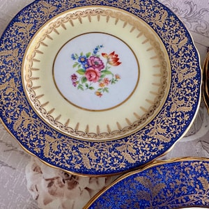 May include: A set of vintage porcelain plates. Each plate features a cream-colored center with a floral bouquet and a gold rim. The outer rim is navy blue with a gold leaf pattern. The plates have a scalloped edge.