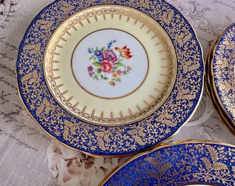 May include: A set of vintage porcelain plates. Each plate features a cream-colored center with a floral bouquet and a gold rim. The outer rim is navy blue with a gold leaf pattern. The plates have a scalloped edge.