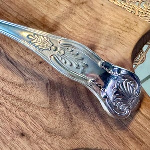 May include: A silver serving utensil with an ornate, detailed handle. The handle has a floral design and is polished to a high shine.
