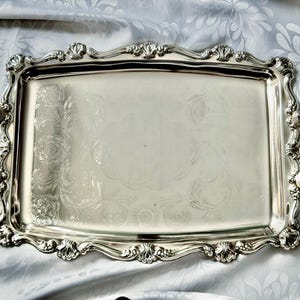 Pu&ograve; includere: Un vassoio da portata rettangolare color argento caratterizzato da volute in rilievo e dettagli a forma di conchiglia lungo i bordi smerlati. Il vassoio include due manici decorativi sui lati e un sottile motivo floreale inciso sulla superficie.