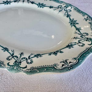 May include: An oval ceramic serving platter with a cream-coloured base and a scalloped edge. The rim features a decorative green transferware pattern consisting of floral garlands, ribbons, and intricate scrollwork borders around the centre.