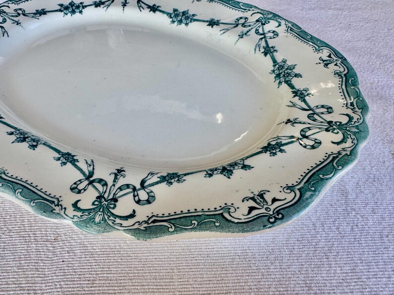 May include: An oval ceramic serving platter with a cream-coloured base and a scalloped edge. The rim features a decorative green transferware pattern consisting of floral garlands, ribbons, and intricate scrollwork borders around the centre.