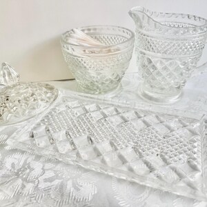 May include: A clear pressed glass vanity set featuring a rectangular tray, a lidded jar, a small bowl, and a creamer pitcher. Each piece displays a consistent diamond-cut pattern with faceted edges, reflecting light across the textured surfaces.