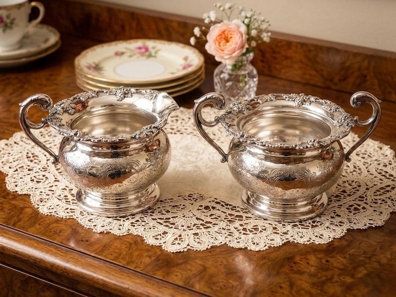 Puede incluir: Dos recipientes de metal plateado con patrones florales en relieve en los bordes y cuerpos. Cada pieza presenta una base redondeada, una abertura ancha y dos asas curvas en los lados opuestos, con un acabado met&aacute;lico pulido.