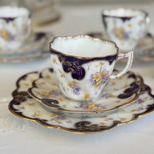 May include: A fine bone china teacup and saucer set. The cup and saucer have a white background with a floral design in yellow and purple, highlighted with navy blue and gold edging. The set is displayed on a white surface.