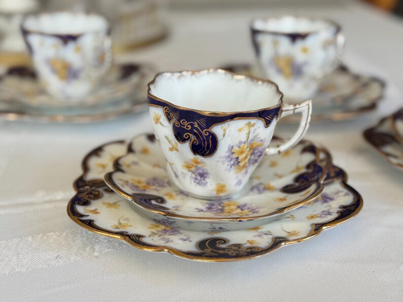 May include: A fine bone china teacup and saucer set. The cup and saucer have a white background with a floral design in yellow and purple, highlighted with navy blue and gold edging. The set is displayed on a white surface.