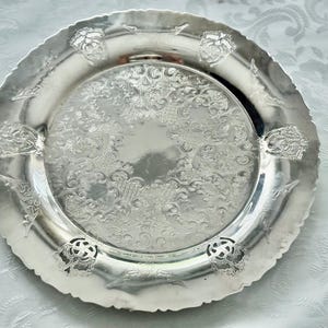 May include: A silver-colored decorative plate with an ornate, embossed floral design. The plate has a scalloped edge and features detailed patterns and floral accents. This vintage-style plate is suitable for serving or display.