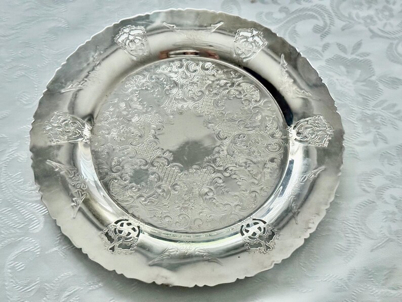 May include: A silver-colored decorative plate with an ornate, embossed floral design. The plate has a scalloped edge and features detailed patterns and floral accents. This vintage-style plate is suitable for serving or display.