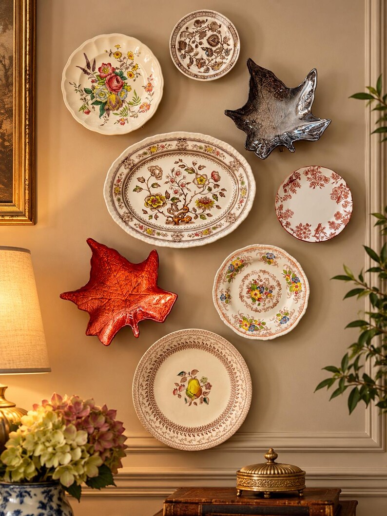 May include: A collection of decorative wall plates and leaf-shaped dishes arranged on a wall. The display includes floral patterned plates, a red leaf dish, a dark metallic leaf dish, and a plate featuring a central pear illustration.