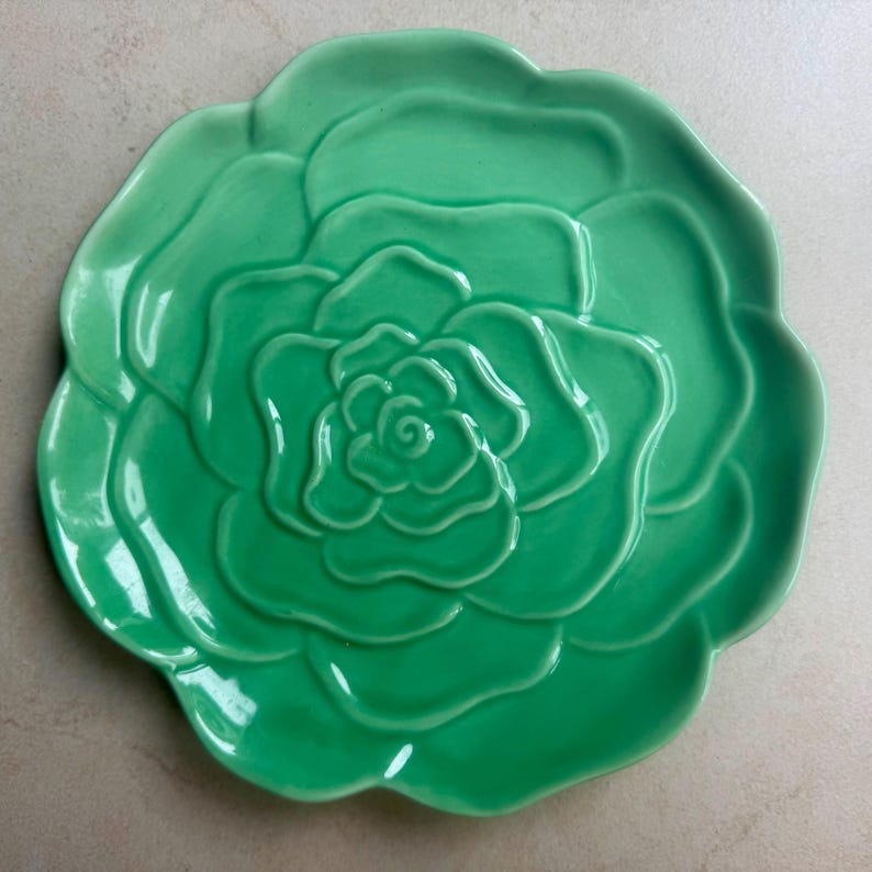 Puede incluir: Un plato de cer&aacute;mica circular en un vibrante color verde menta, que presenta un dise&ntilde;o en relieve tridimensional de una flor en flor con p&eacute;talos curvos y superpuestos que irradian desde el centro hacia los bordes ondulados y festoneados.