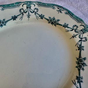 May include: An oval ceramic platter with a cream-coloured base and a scalloped edge. The rim features a dark teal transferware pattern consisting of delicate floral garlands, small blossoms, and decorative ribbons draped around the border.