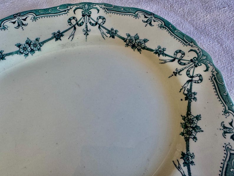May include: An oval ceramic platter with a cream-coloured base and a scalloped edge. The rim features a dark teal transferware pattern consisting of delicate floral garlands, small blossoms, and decorative ribbons draped around the border.