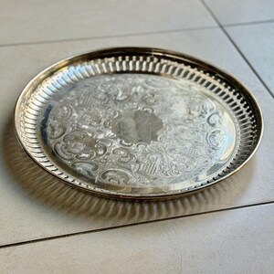 Puede incluir: Una bandeja ovalada plateada con intrincados dise&ntilde;os florales y de volutas. La bandeja tiene un borde decorativo calado y un borde elevado. La superficie reflectante y el dise&ntilde;o sugieren un estilo vintage o antiguo.