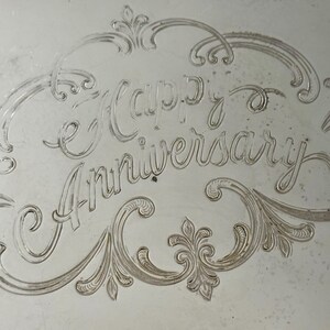 May include: A silver-toned metallic surface featuring an engraved inscription that reads Happy Anniversary. The text is centered within an ornate, swirling decorative border with intricate line work and symmetrical flourishes.