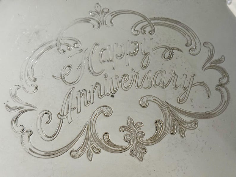 May include: A silver-toned metallic surface featuring an engraved inscription that reads Happy Anniversary. The text is centered within an ornate, swirling decorative border with intricate line work and symmetrical flourishes.