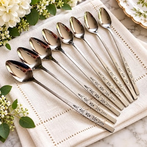 May include: A set of seven silver-toned metal spoons arranged in a diagonal row. Each spoon features a long, slender handle decorated with a repeating floral and scroll pattern. The polished bowls of the spoons reflect light, showing a smooth finish.