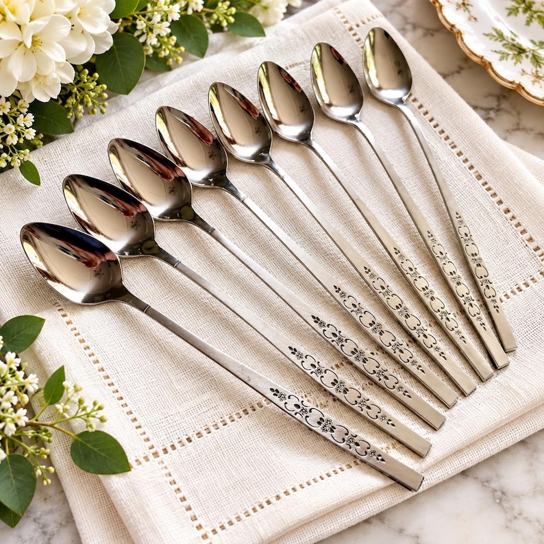 May include: A set of seven silver-toned metal spoons arranged in a diagonal row. Each spoon features a long, slender handle decorated with a repeating floral and scroll pattern. The polished bowls of the spoons reflect light, showing a smooth finish.