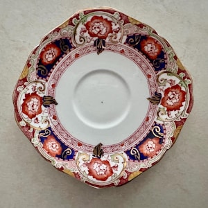 May include: A round ceramic saucer with a scalloped edge featuring a white center and a decorative border. The border displays a pattern of red floral motifs, deep blue panels, and gold scrollwork accents arranged in a symmetrical design.