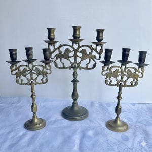 Pu&ograve; includere: Tre candelabri antichi in ottone. Ogni supporto ha pi&ugrave; bracci per candele ed &egrave; decorato con un motivo a leone. I candelabri variano nelle dimensioni e hanno un'estetica vintage.