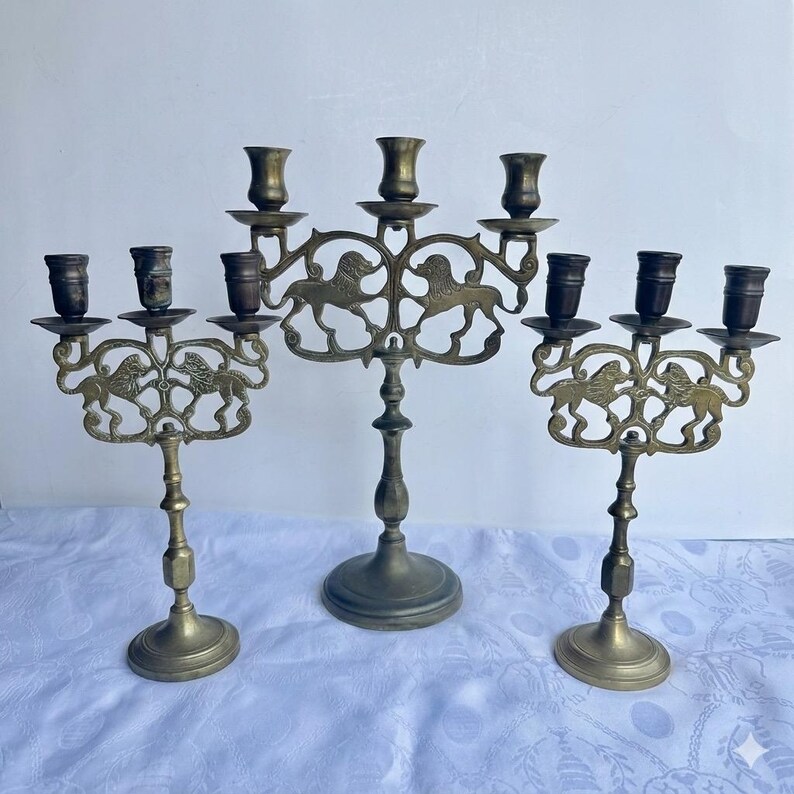 Pu&ograve; includere: Tre candelabri antichi in ottone. Ogni supporto ha pi&ugrave; bracci per candele ed &egrave; decorato con un motivo a leone. I candelabri variano nelle dimensioni e hanno un'estetica vintage.