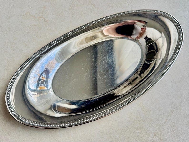 K&ouml;nnte beinhalten: Ein ovales Serviertablett aus silberfarbenem Metall mit einer polierten, reflektierenden Oberfl&auml;che. Der Rand des Tabletts ist mit einem dekorativen Perlenmuster versehen, das entlang des gesamten Umfangs verl&auml;uft und eine texturierte Kante bildet.