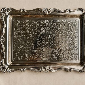 Puede incluir: Una bandeja rectangular de metal plateado que presenta un intrincado patr&oacute;n de volutas en relieve en la superficie central. La bandeja tiene un borde elevado y ornamentado con bordes festoneados y tallas detalladas.
