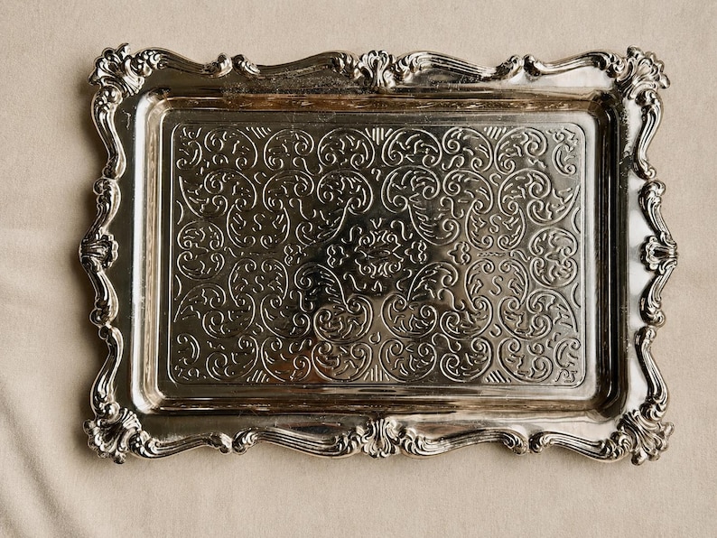 Puede incluir: Una bandeja rectangular de metal plateado que presenta un intrincado patr&oacute;n de volutas en relieve en la superficie central. La bandeja tiene un borde elevado y ornamentado con bordes festoneados y tallas detalladas.